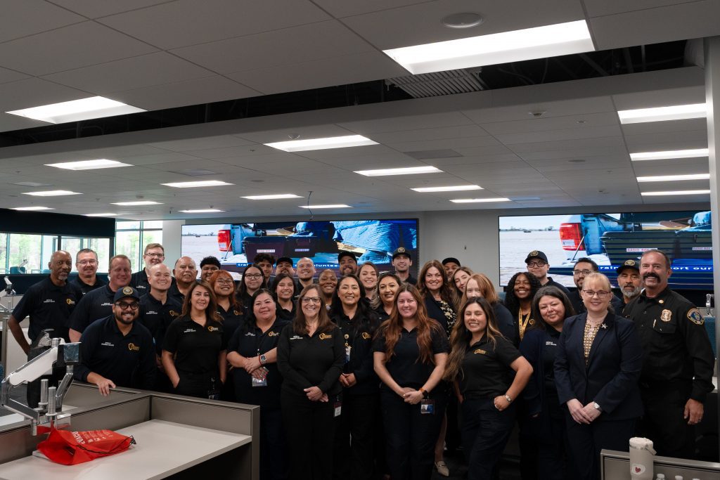 Group photo of San Bernardino County Office of Emergency Services staff, public safety partners, and county leadership inside the Emergency Operations Center at the Valley Communications Center. Team members stand together in front of large display screens and workstations.