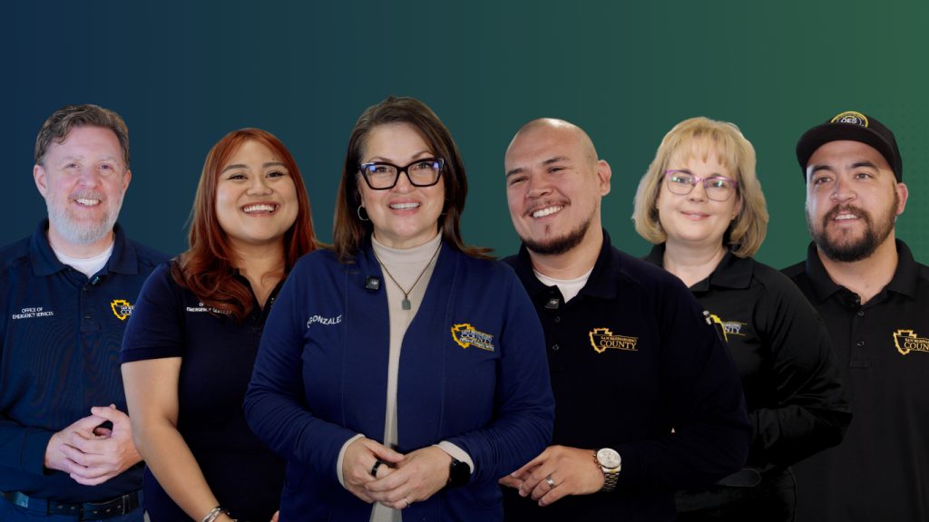 Group photo of six San Bernardino County Office of Emergency Services team members standing together and smiling against a blue and green gradient background. The employees wear Office of Emergency Services or San Bernardino County branded apparel, including polos, jackets and a cap, representing the diverse team that supports emergency management efforts before, during and after emergencies.