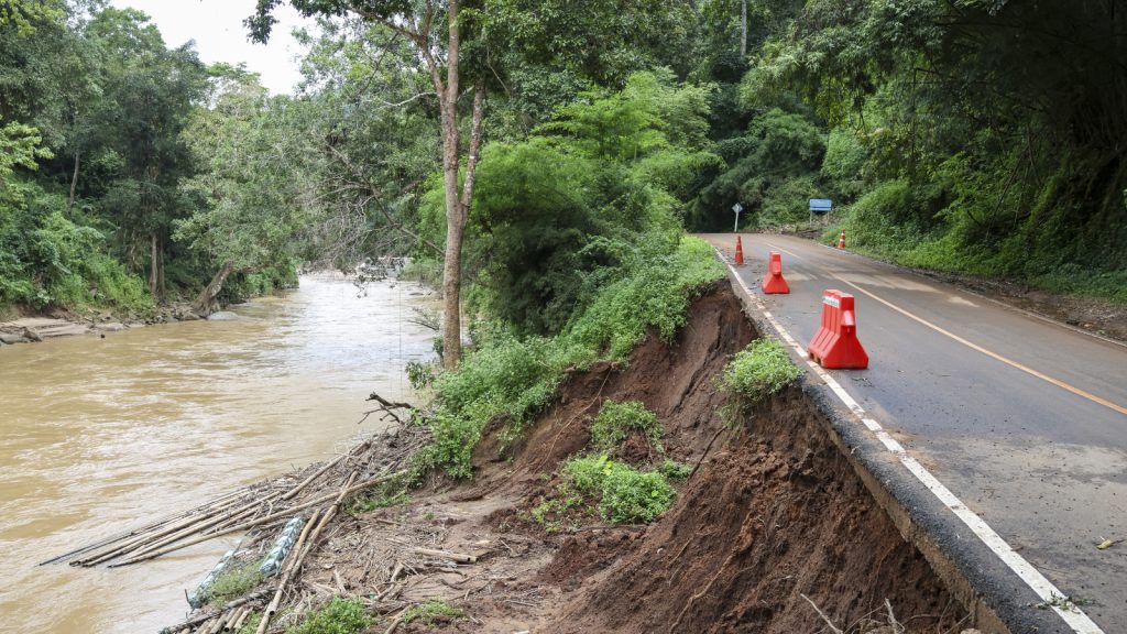 A rain-swollen river runs alongside a damaged roadway where erosion has washed away part of the embankment. Orange traffic barriers mark the affected section of road surrounded by dense vegetation.